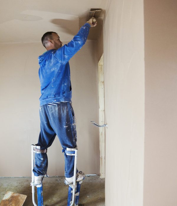 A plasterer wearing stilts smoothing fresh plaster high up on the walls of a house under construction.